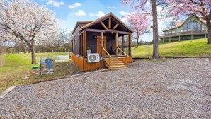 A small wooden cabin with a porch, a gravel driveway, and a pond in the background. - Romantic Tiny Home with Pond, Pier & Firepit (Denison)