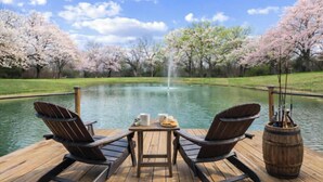 A wooden deck with two Adirondack chairs, a small table with a pitcher, a plate of cookies, and a fishing rod holder. - Romantic Tiny Home with Pond, Pier & Firepit (Denison)