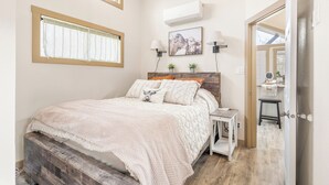 A bedroom with a bed, bedside tables, a window, an air conditioner, and a door leading to another room. - Romantic Tiny Home with Pond, Pier & Firepit (Denison)