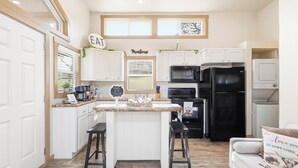 A modern kitchen with white cabinets, a black refrigerator, a microwave, and a coffee maker. There is a sign that says "Farmhouse" and another that says "Eat." - Romantic Tiny Home with Pond, Pier & Firepit (Denison)