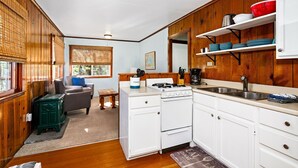 Una cocina compacta con armarios blancos, una estufa y un lavamanos. Hay una zona de comedor con una mesa y sillas, y una ventana con persianas.