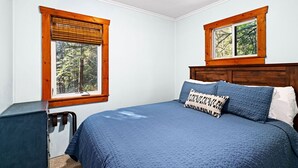 Un dormitorio con cama, dos ventanas de madera y vista a árboles.