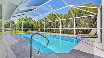 A swimming pool with a glass enclosure, surrounded by a patio area with chairs and a view of palm trees.