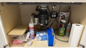 Ein Küchenspüle mit Filtersystem, ein Mülleimer, eine Flasche Desinfektionsmittel, ein Müllbeutelspender und eine Toilettenpapierrolle.