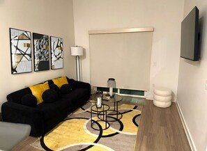 A modern living room with a black sofa, yellow cushions, abstract wall art, a glass coffee table, and a flat-screen TV mounted on the wall.