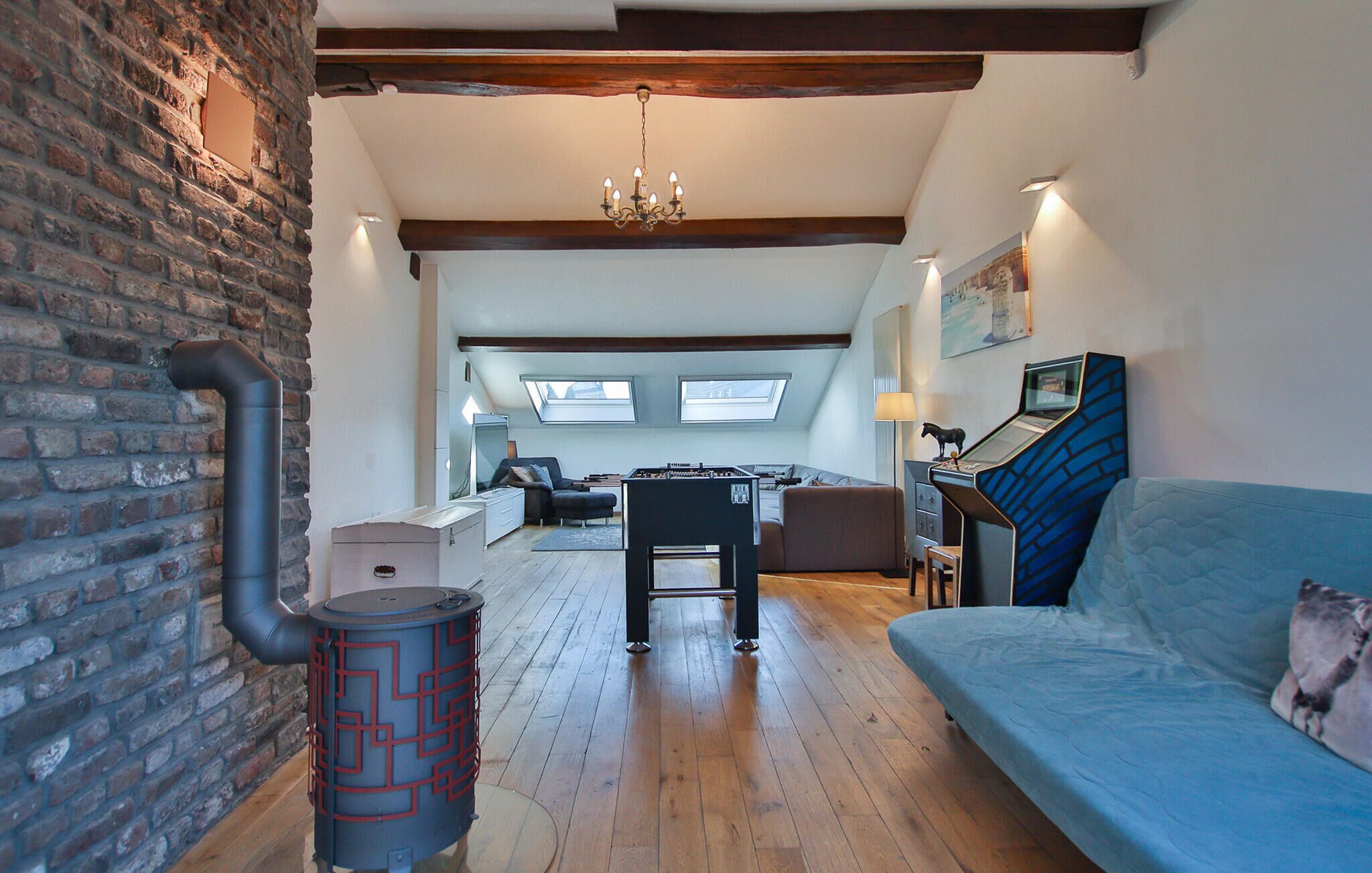 A loft-style living area with exposed brick walls, a wooden floor, a blue sofa, a foosball table, and a large window providing natural light.