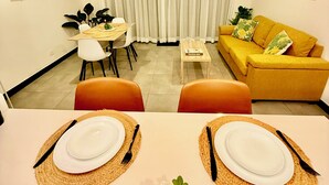 A dining area with a table set for two, a sofa, a TV mounted on the wall, and a window with curtains.