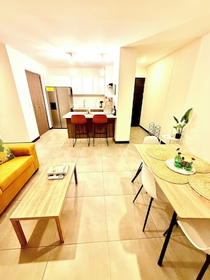 Un salon moderne avec un canapé, une table basse et une table à manger. On aperçoit à l’arrière-plan un coin cuisine équipé d’un réfrigérateur, d’une cuisinière et de tabourets de bar.