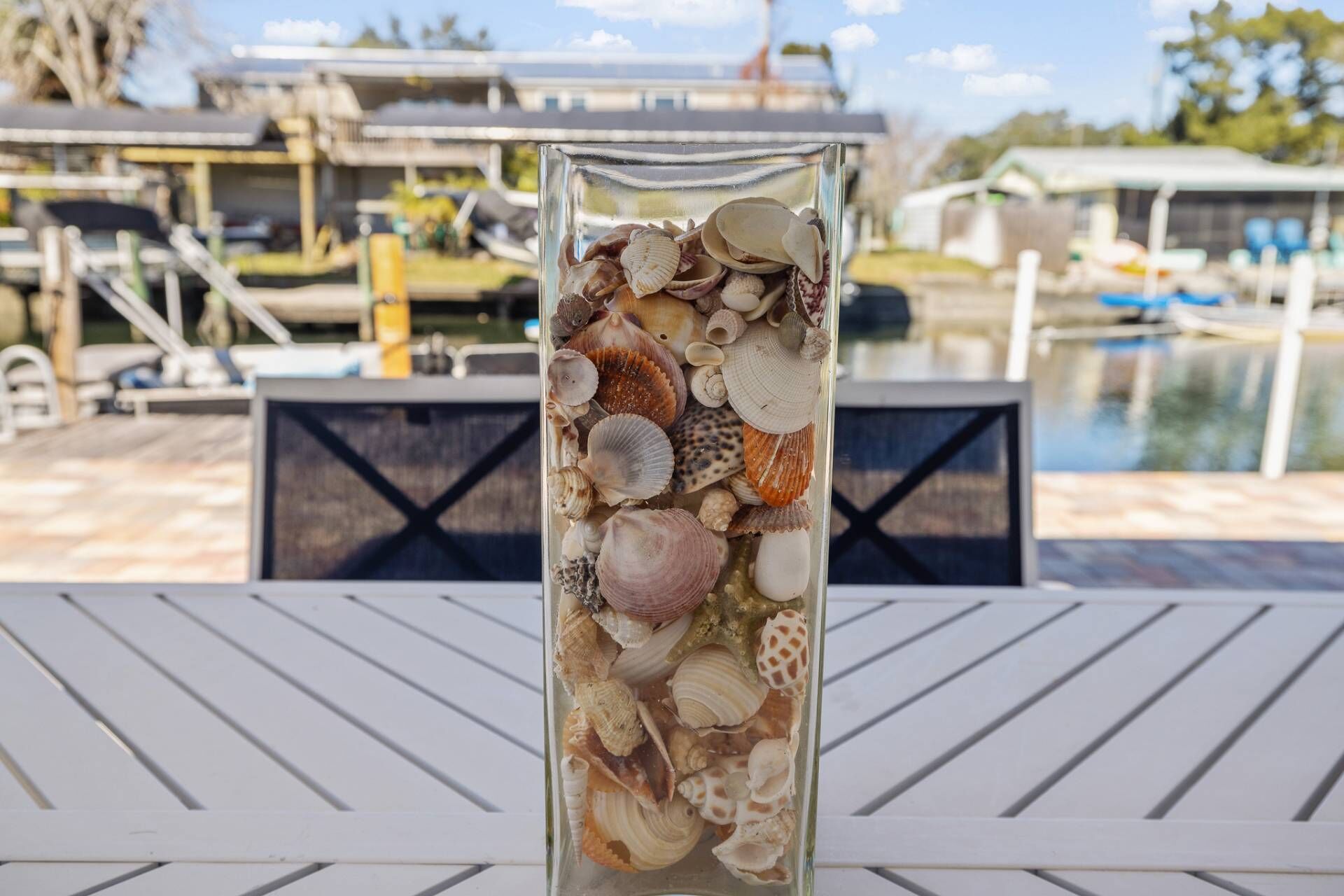 A glass vase filled with various seashells placed on a white table with a view of a pool and outdoor seating area.