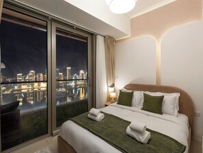 A hotel room with a large window offering a nighttime city view, a neatly made bed with green and white bedding, and a bedside lamp. - Charming Flat with Sea & AIN View (Dubai)