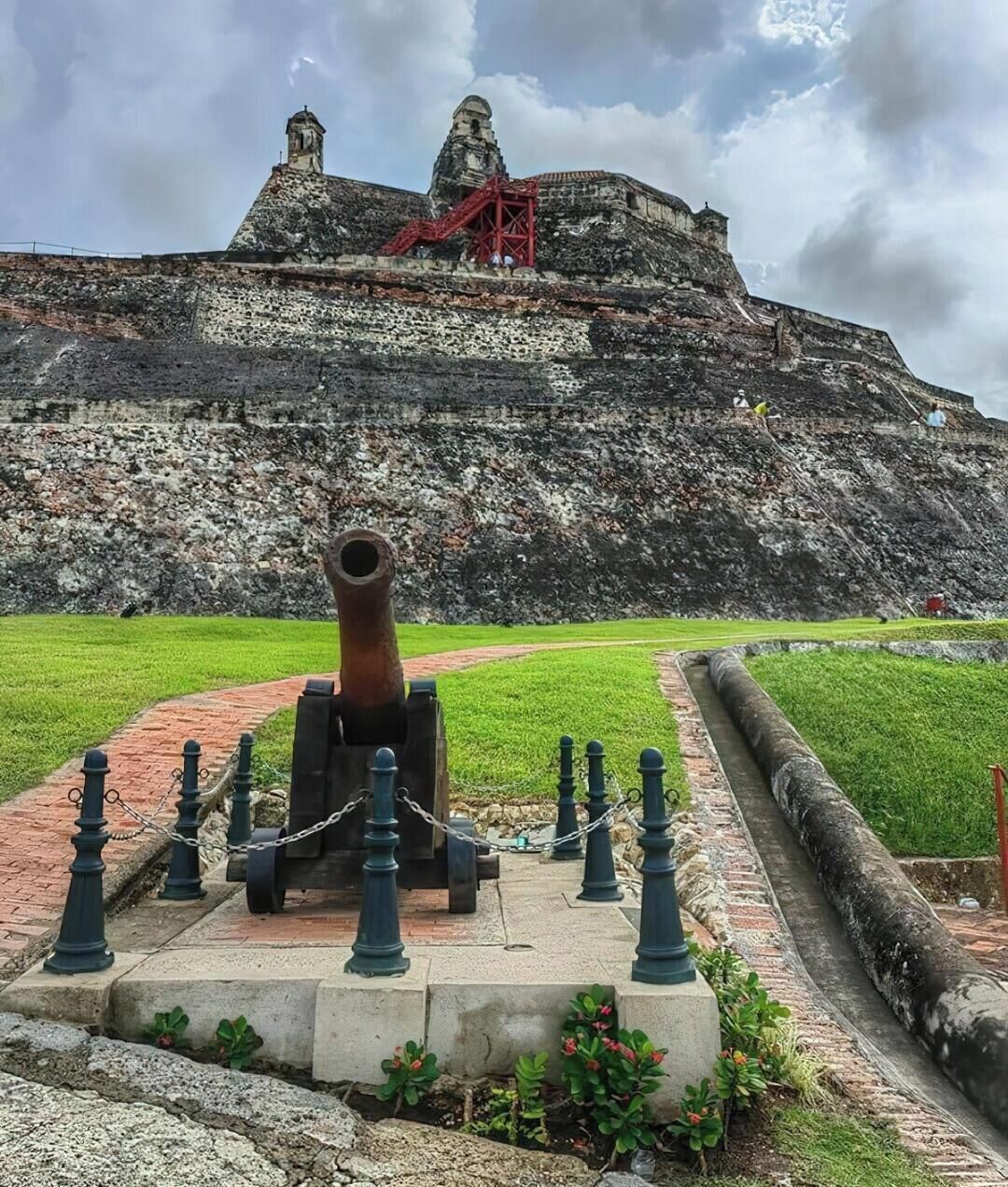A historical fort with a large cannon in the foreground, a red staircase, and a grassy area.
