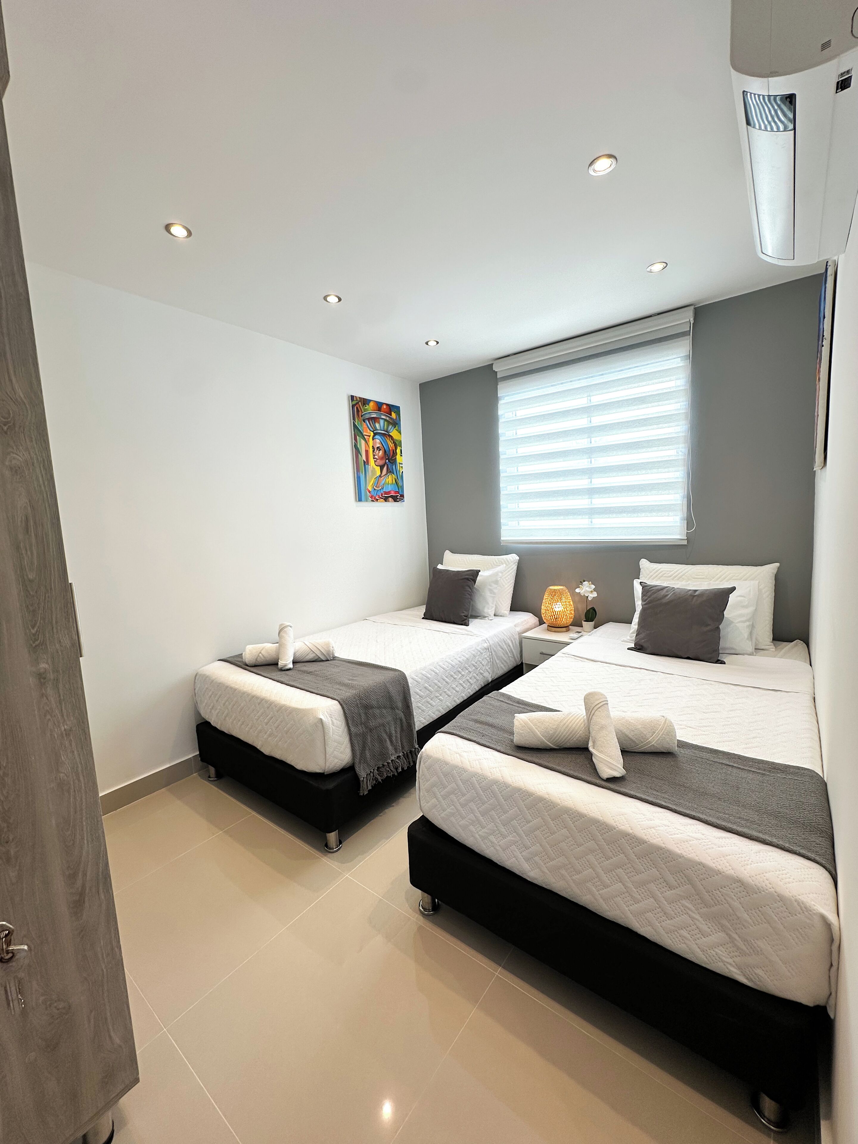 A modern hotel room with two beds, a large window with blinds, a painting on the wall, and a ceiling air conditioner.