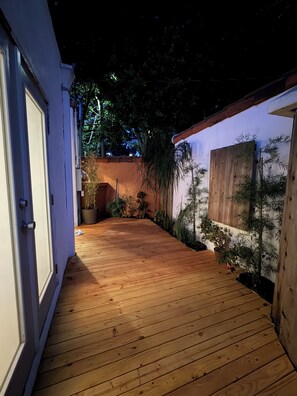 Eine Holzterrasse führt zu einem Gebäude mit einer weißen Tür, umgeben von Topfpflanzen und einem Holzzaun.