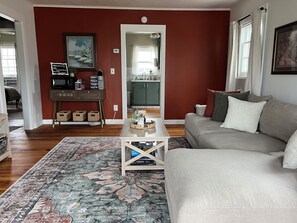 A living room with a sofa, a coffee table, a rug, a console table with decorative items, and a view into the kitchen.