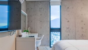 Een moderne slaapkamer met een groot bed, een aan de muur bevestigde televisie, een klein bureau met stoel, en een raam met uitzicht over de stad.