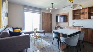 A modern living room with a sofa, coffee table, and dining area with chairs. There is a kitchen with a TV and wooden cabinets.