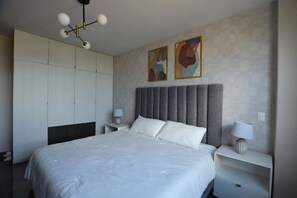 Un dormitorio moderno con una cama grande, un cabecero tapizado en gris y dos mesitas de noche con lámparas.