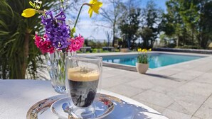 A glass of iced coffee on a white plate with a spoon, placed on a table outdoors with a pool and flowers in the background. - Suite Ravenna : Charm, Cathedral height & Luxury, Swimming pool - Domaine de Pagnon (Saint-Gervais)