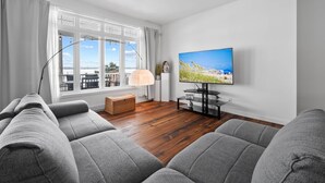 Un salon moderne avec des canapés gris, un téléviseur fixé au mur, une grande fenêtre avec des rideaux et un plancher en bois.