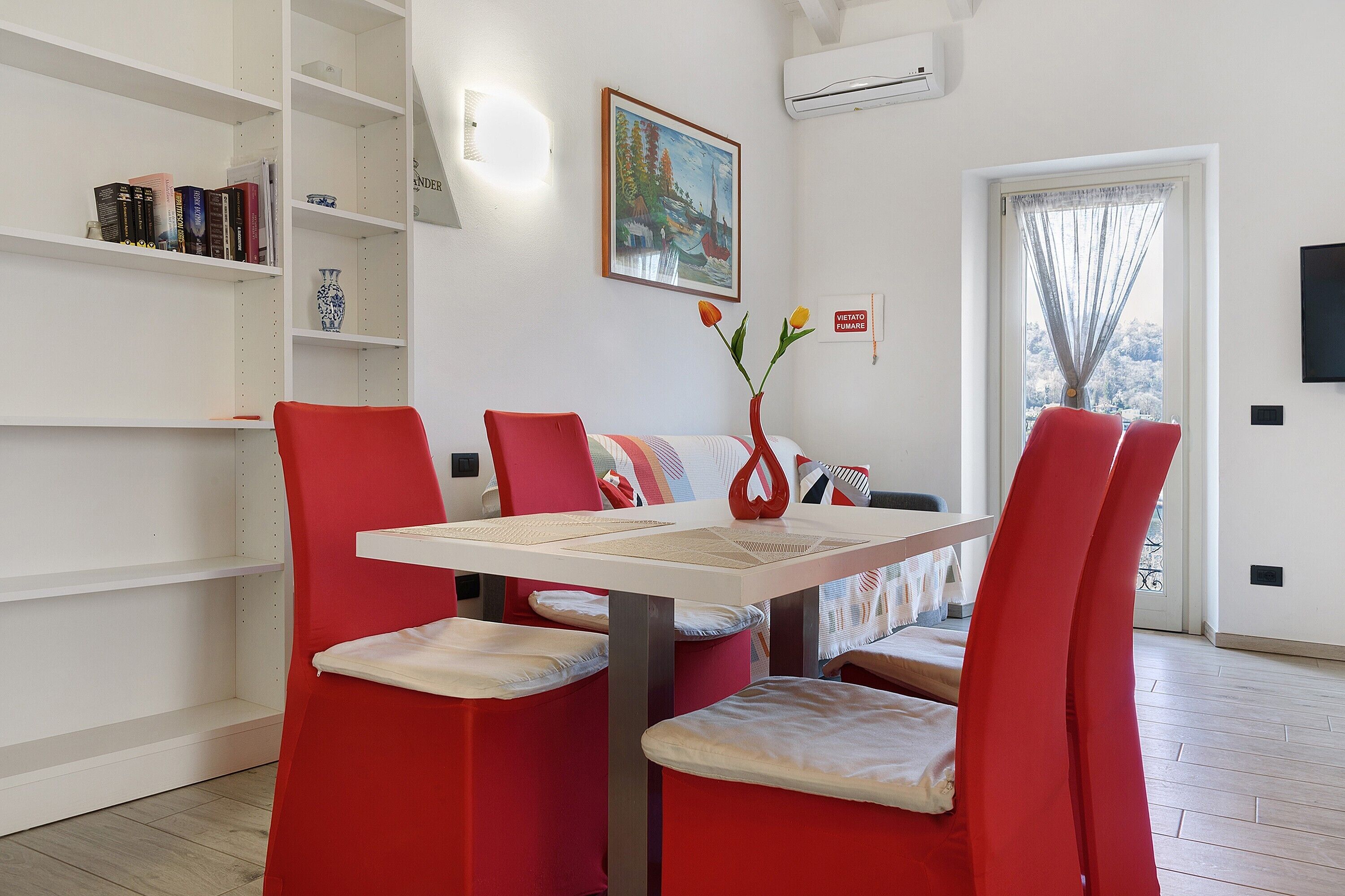 Un coin repas avec une table blanche, des chaises rouges, un vase avec des fleurs et une bibliothèque contenant des livres et un autre vase.