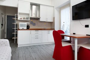 Eine kompakte Küche mit weißen Schränken, einer Mikrowelle und einer Kaffeemaschine. Ein roter Stuhl und ein kleiner Tisch, an der Wand ist ein Fernseher angebracht.