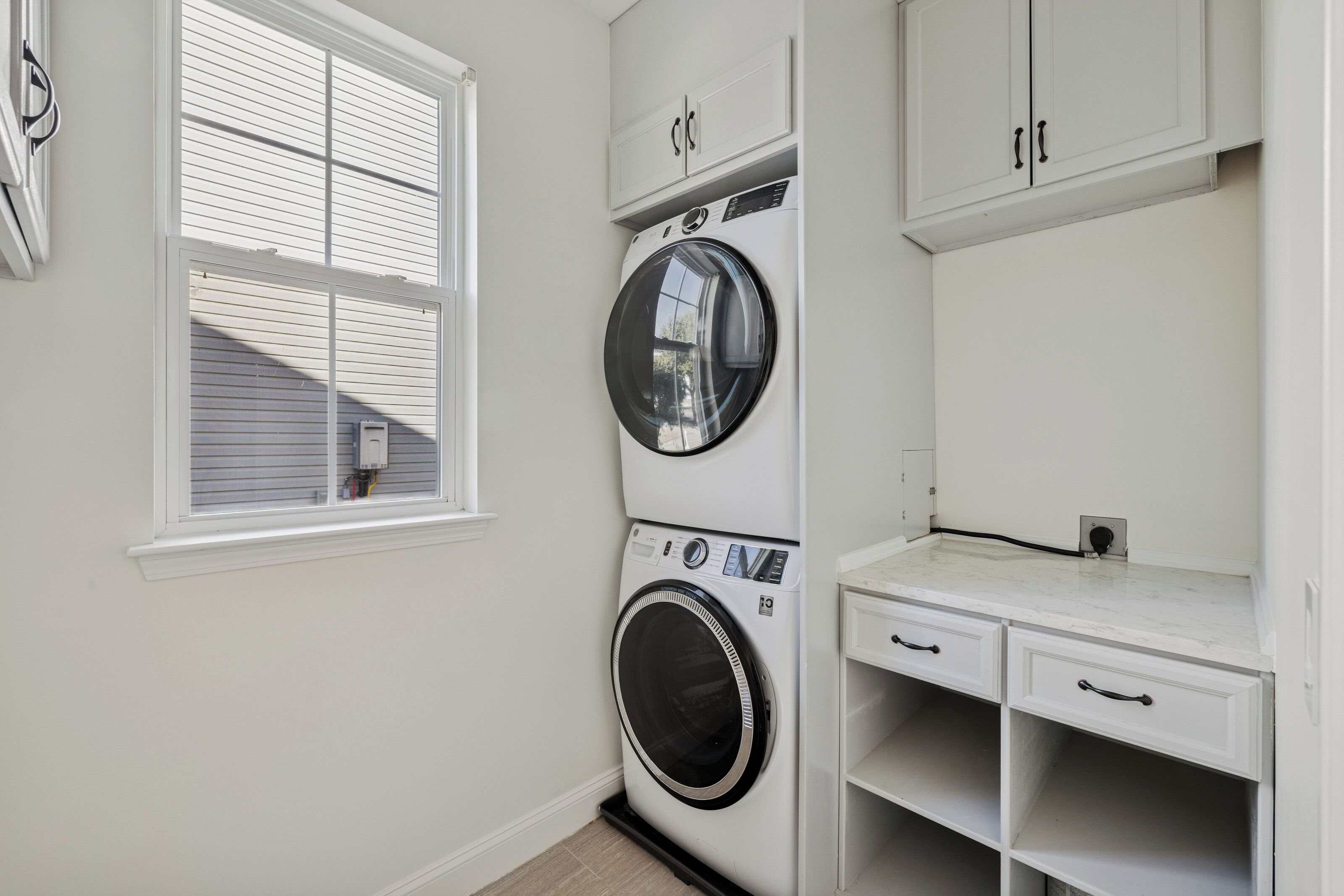 Une buanderie équipée d’un lave-linge et d’un sèche-linge à chargement frontal, d’une fenêtre avec des stores et d’une petite étagère avec des tiroirs.