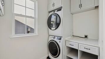 Une buanderie équipée d’un lave-linge et d’un sèche-linge à chargement frontal, d’une fenêtre avec des stores et d’une petite étagère avec des tiroirs.