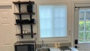 A compact kitchen with a built-in microwave, a sink, and a refrigerator. There is a window with blinds, a door, and a shelf unit with kitchen items. - Amazing Studio in Pasadena! (Pasadena)