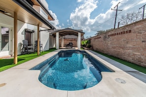 Una piscina rectangular con jacuzzi integrado, rodeada por un muro de ladrillo y un patio cubierto con sillas.