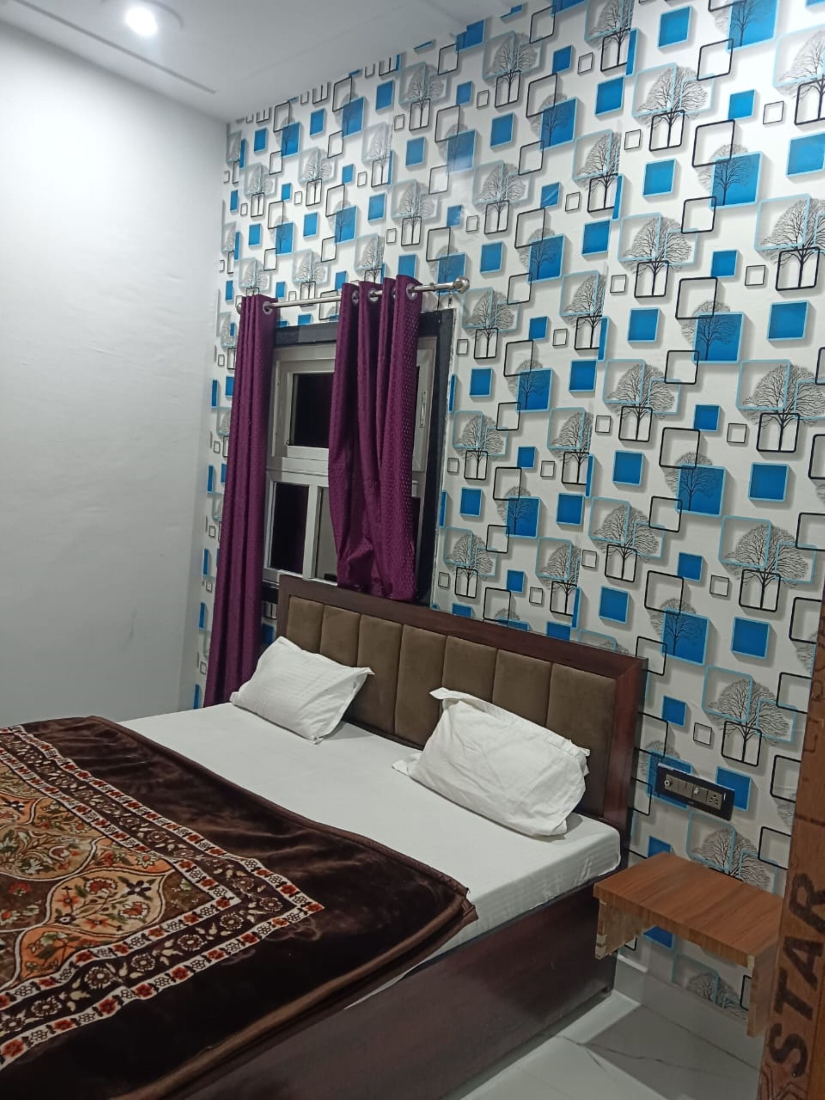 Ein Schlafzimmer mit einem Bett, einem Fenster mit violetten Vorhängen, einer Wand mit Muster und einem kleinen Holztisch.