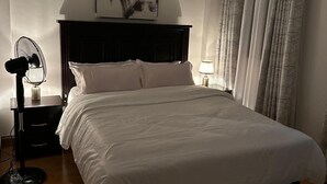 Een slaapkamer met een bed, nachtkastje met lamp, ventilator en een kunstwerk aan de muur.