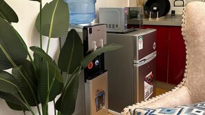 Een compacte keuken met een waterdispenser, magnetron en koelkast. Er staat een potplant en er is een gestreept fauteuil.