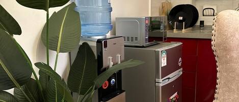 A compact kitchen with a water dispenser, microwave, and refrigerator. There is a potted plant and a patterned armchair.