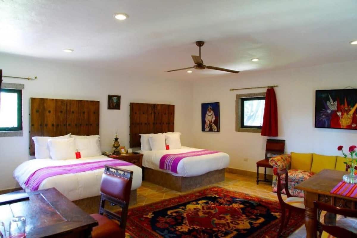 Habitación de hotel con dos camas, cabecero de madera, ventilador de techo, mesa de comedor con sillas y una alfombra colorida.