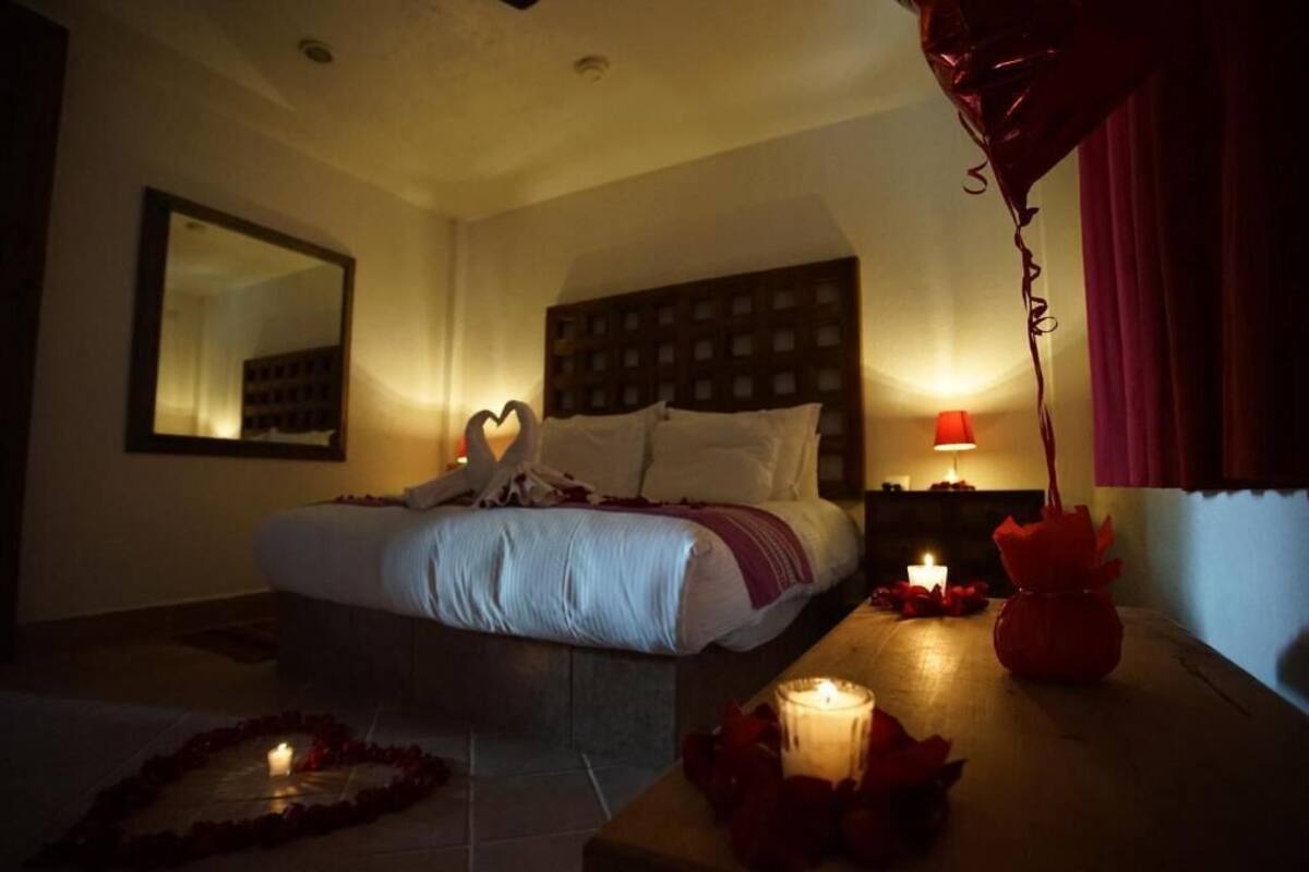Un habitación de hotel con poca luz, con una cama, un espejo, una mesita de noche con una lámpara, y un arreglo decorativo de pétalos de rosa y velas con forma de corazón.