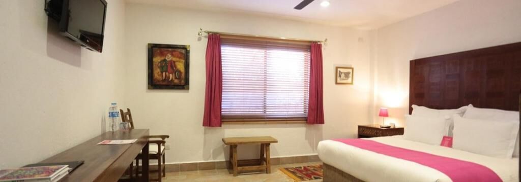 A hotel room with a bed, a desk, a chair, a window with curtains, a television, and a painting on the wall.
