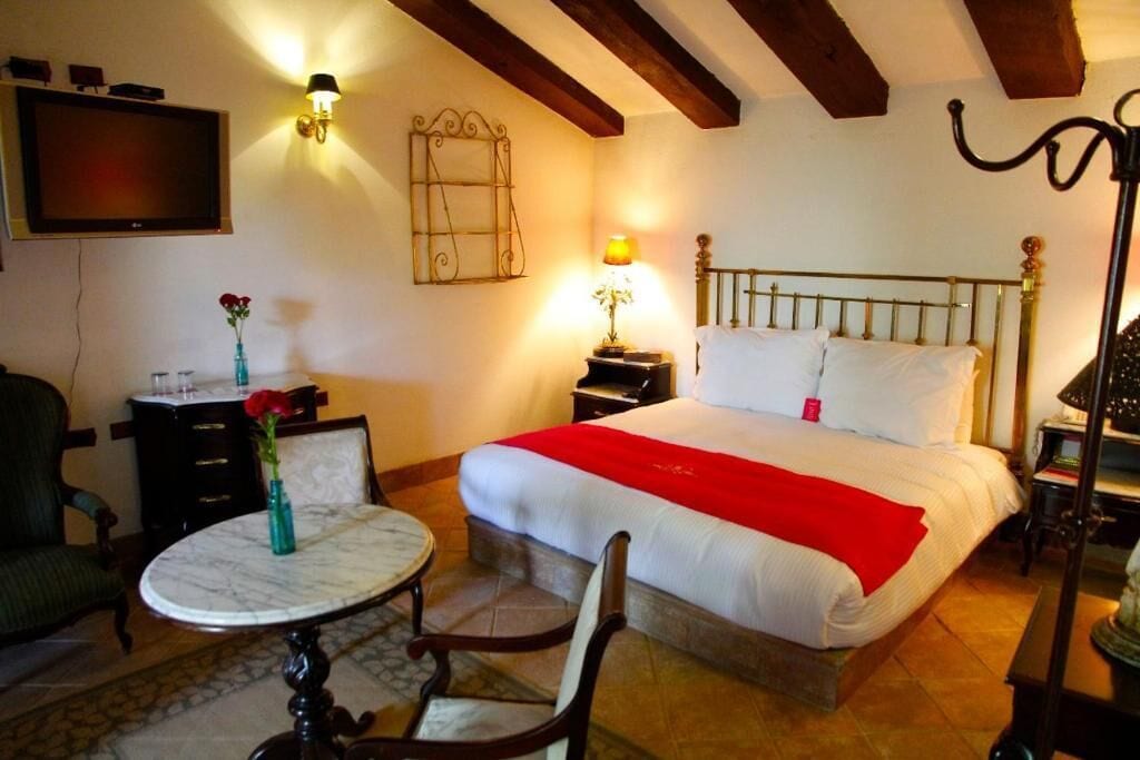 Un dormitorio con cama, mesitas de noche, televisor, lámpara, silla y una pequeña mesa.