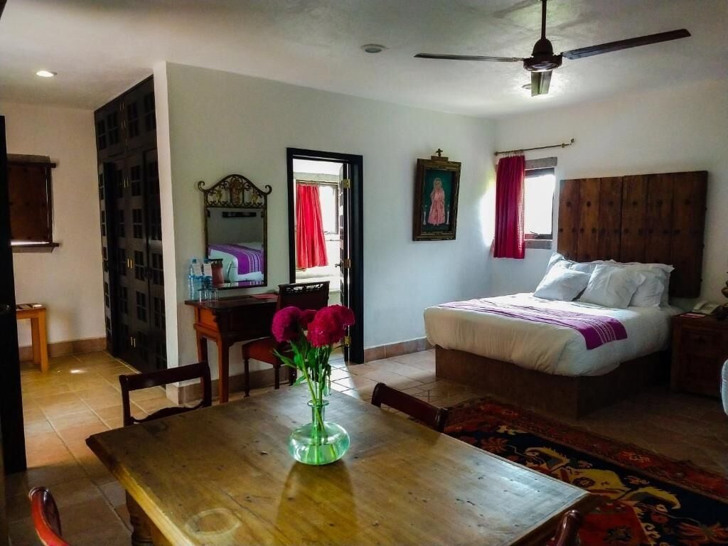 Un dormitorio con una cama, una mesa de madera con un florero, un escritorio con un espejo y un ventilador de techo.