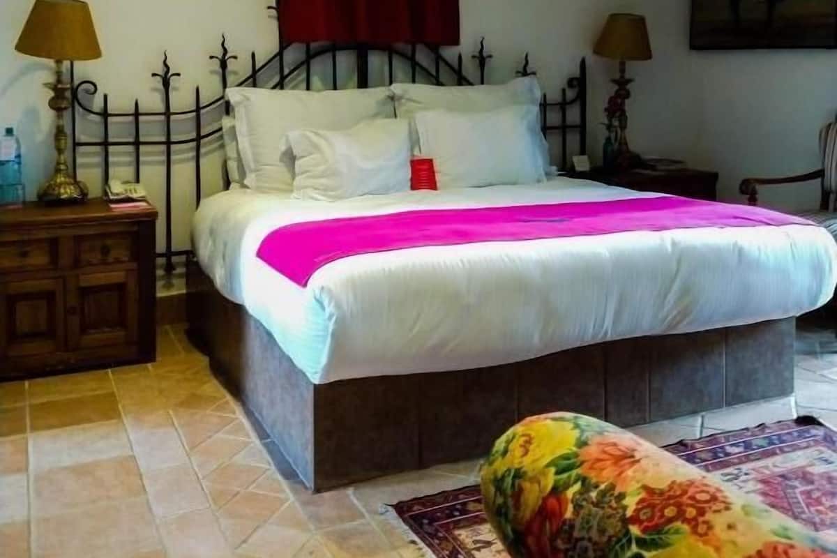 Un dormitorio con una cama, dos lámparas de noche, un ventilador de techo, una cortina roja, un cuadro y un reposapiés estampado con motivos florales.