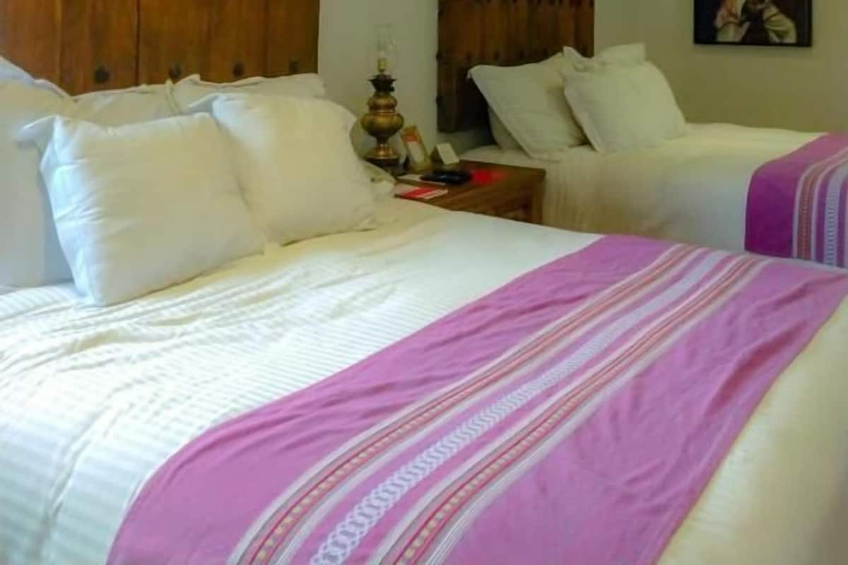 Habitación de hotel con dos camas, cabeceros de madera, ropa blanca y una mesita con lámpara.
