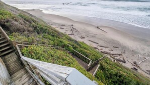 Eine Holztreppe führt hinunter zu einem Strand, im Hintergrund sind Wellen zu sehen.