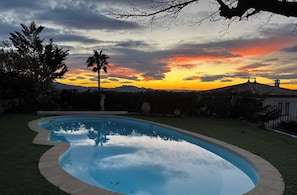 Une piscine à bord incurvé, entourée d’une pelouse verdoyante, avec en arrière-plan un coucher de soleil sur les montagnes.