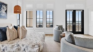 A spacious room with a large bed, a grey armchair, and a view of the ocean through large windows. - Driftwood Waves 116 LES (Port Aransas)