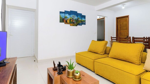 A living room with a yellow sofa, a coffee table with decorative items, a flat-screen TV, and a wall-mounted painting.