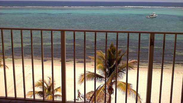 Blick auf einen Strand vom Balkon aus, mit Geländer, Palmen und einem Boot im Wasser.