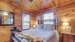 Un dormitorio de cabaña de madera con cama, mesita de noche, lámpara y un osito de peluche en el cabecero.