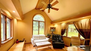 Ein geräumiges Zimmer mit hoher Decke, einem Deckenventilator, einem großen Fenster mit Blick auf Grünflächen, einem weißen Sofa, einem schwarzen Ledersessel und einem Holztisch mit Computerarbeitsplatz.