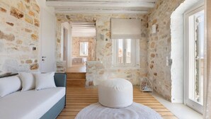 Ein Zimmer mit Steinwänden und Holzboden, einem weißen Sofa, einem runden weißen Hocker und einem Fenster mit Aussicht.