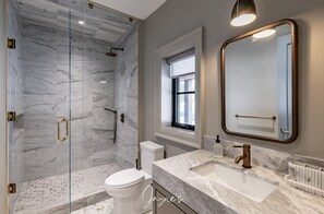 Et moderne badeværelse med en brusekabine i glas, et hvidt toilet, en marmorvask og et stort spejl.