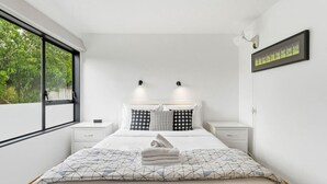 Een moderne slaapkamer met een groot bed, twee nachtkastjes, een raam met uitzicht op groen en een ingelijst schilderij aan de muur.